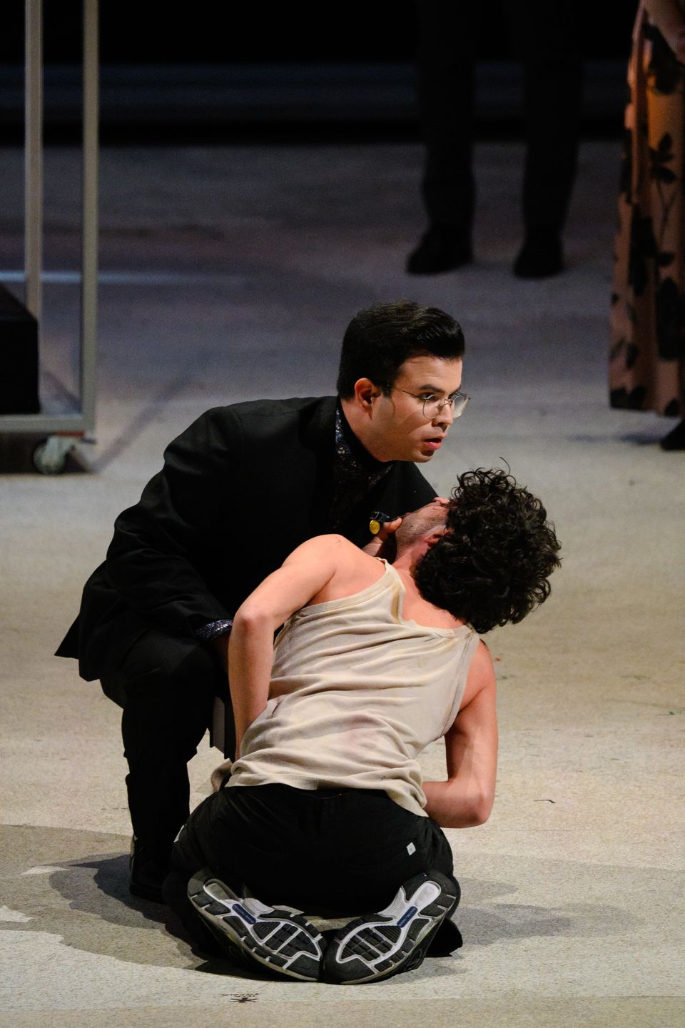 A performer in a dark suit and glasses supports another who is kneeling and leaning backwards on stage.