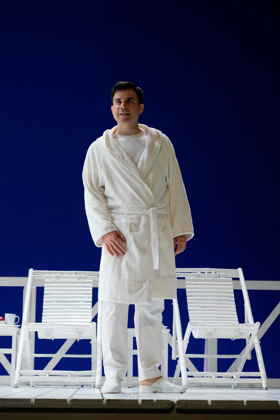 Enrico Basso in a white bathrobe on stage, in front of two white deckchairs and a blue backdrop.
