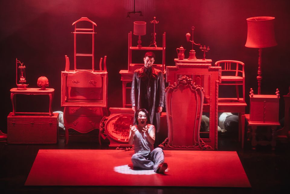 A red-lit stage: a performer in a dark coat stands behind another seated on the floor, among stacked furniture, frames and lamps.