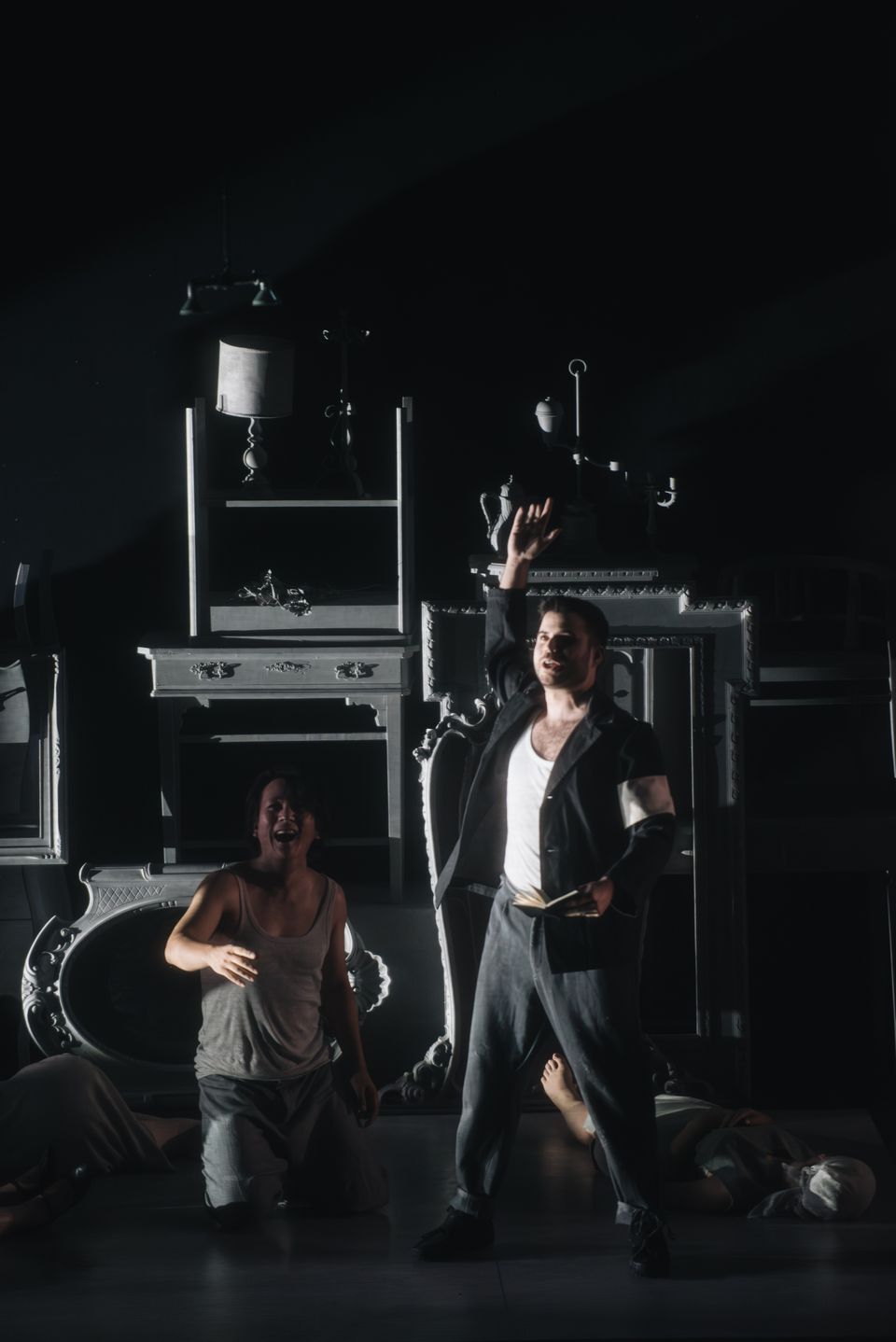 A performer in a dark jacket raises one hand and holds an open book beside another kneeling performer; stacked frames and furniture fill the stage.