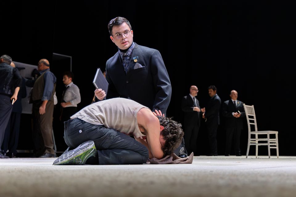A performer in a dark suit and glasses holds a book beside another curled up on the floor; other performers stand in the background.