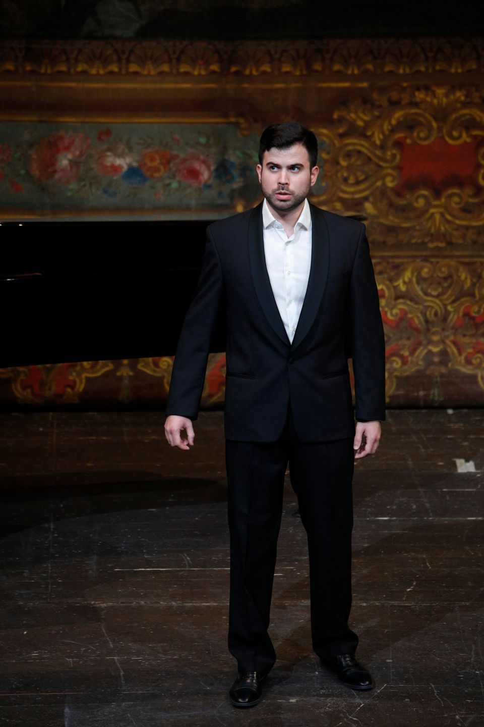 Enrico Basso in a dark suit and white shirt beside a piano, in front of a wall decorated with floral and gold motifs.